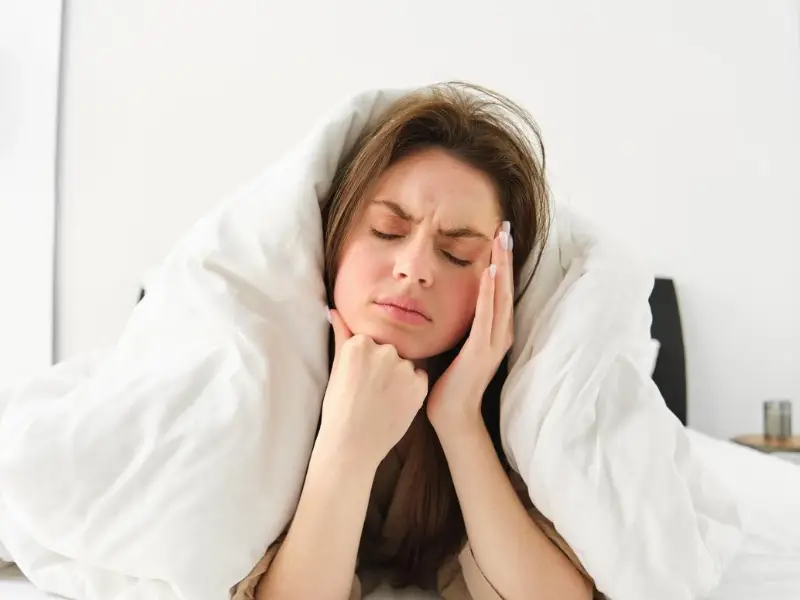 Portrait of woman rolled in blankets in bed frowning and feeling unwell has hangover headache in