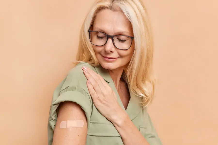 Sideways shot of adult woman with fair hair looks attentively at arm with plaster after vaccine injection protects herself from coronavirus wears spectacles and dress isolated over beige wall