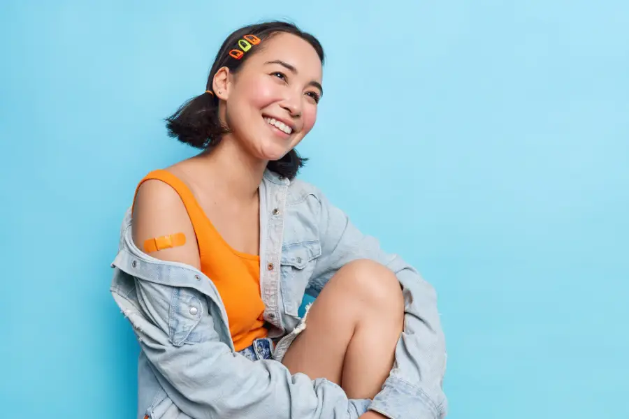 Positive teenage Asian girl with two pony tails looks gladfully aside shows plastered shoulder happy after getting vaccination feels protected wears stylish denim jacket isolated on blue wall